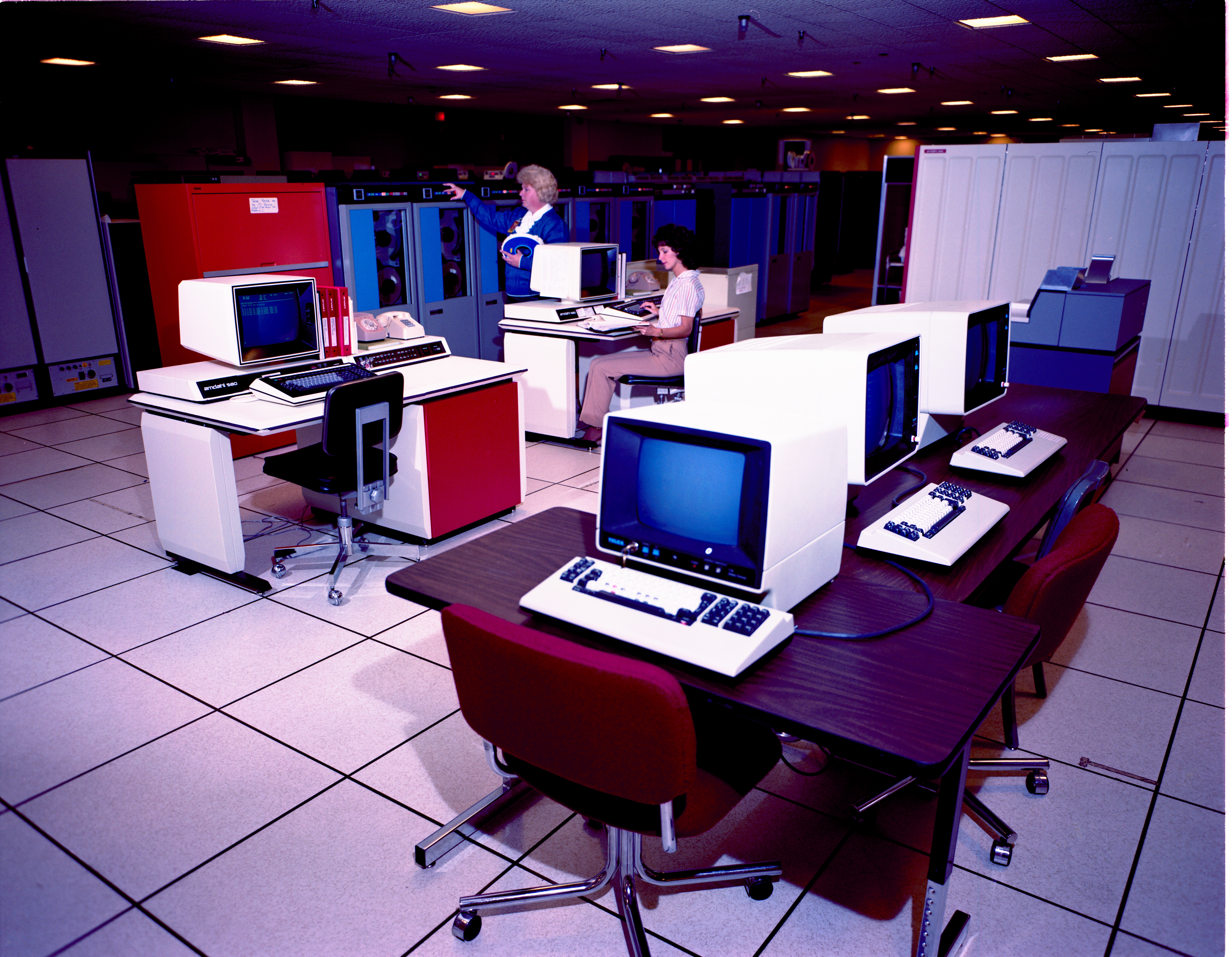 A room with several computers on desks. One woman uses a computer and another woman holds a large reel of magnetic tape.